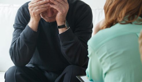 Consultation chez un psychiatre Un homme d'âge mûr, assis dans le cabinet d'un psychiatre, se tenant la tête dans les mains en signe de détresse. En face de lui, un professionnel de santé mentale prend des notes, symbolisant l'écoute et le soutien psychologique.