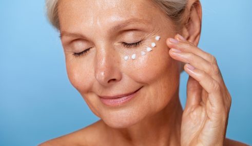 Femme mature souriante, les yeux fermés, appliquant une crème hydratante sous les yeux, illustrant une routine de soin apaisante sur fond bleu Femme mature souriante, les yeux fermés, appliquant une crème hydratante sous les yeux, illustrant une routine de soin apaisante sur fond bleu